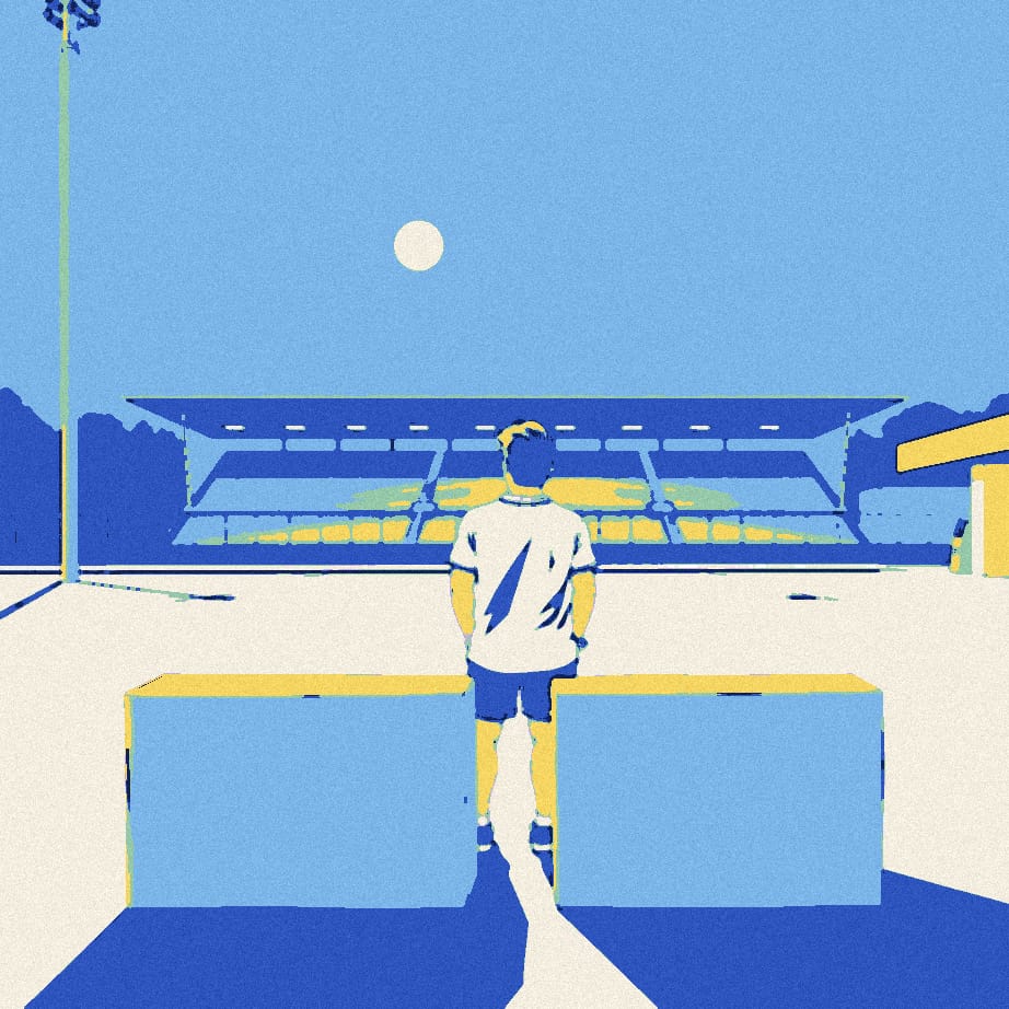 A tiny fan in a plain white shirt stands between two huge bollards on a night-time forecourt, glancing back over his shoulder.