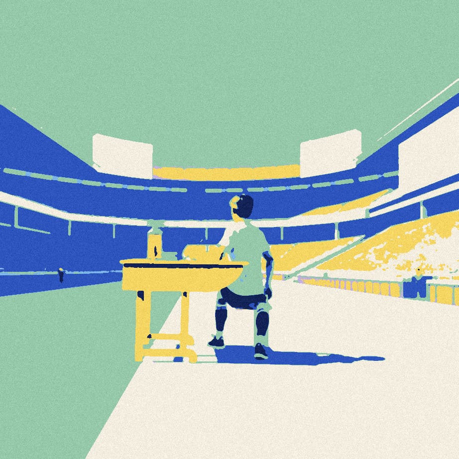 A tiny supporter in a white shirt sits at a giant school desk with a flask on an empty stadium forecourt on a sunny day.