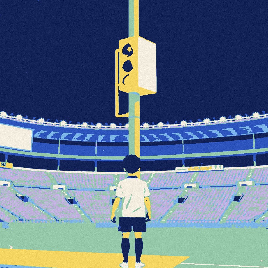 A tiny footballer seen from behind waits at a towering pedestrian crossing signal outside a vast empty stadium at dusk.