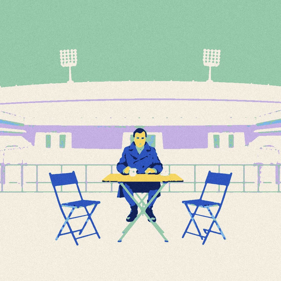 A man in a coat sits at a small table on an empty stadium forecourt, flanked by two empty folding chairs, each with a cup of tea in front of it
