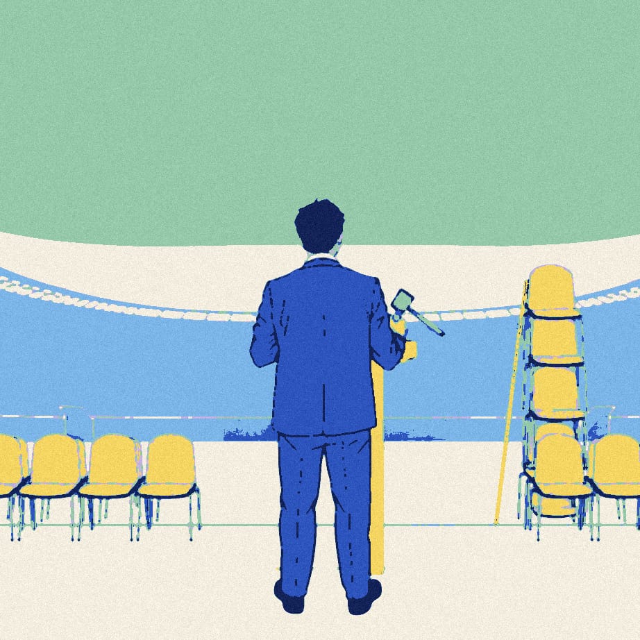 A man seen from behind addresses an empty stadium forecourt from a small lectern next to a towering stack of empty chairs.